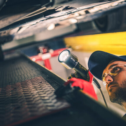 Mechanic Checking Car Undercarriage During Regular Vehicle Inspe Professional Mechanic Checking Car Undercarriage with a Flashlight During Scheduled Vehicle Inspection Appointment at Automobile Station.